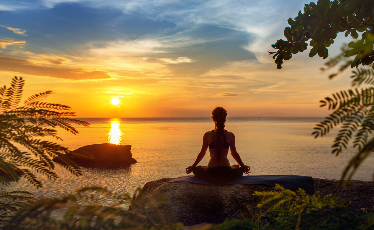 Larimar Properties: A woman is meditating on a rock facing on ocean sunset. The orange sun is reflecting on the ocean.